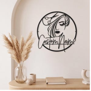 May include: A circular black metal wall sign featuring a stylised silhouette of a person with long hair and a pair of scissors above the head. The text "Custom Name" is written in a cursive script across the lower portion of the circular frame.
