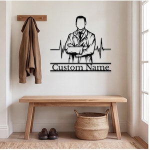May include: A black metal wall art piece featuring a stylized illustration of a doctor with arms crossed, integrated with a horizontal heartbeat EKG line. Below the figure, a rectangular frame contains the black text Custom Name.