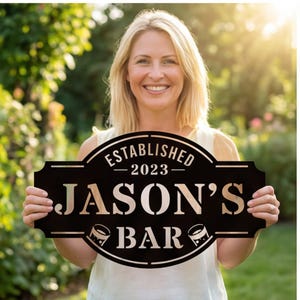 Könnte beinhalten: Ein schwarzes Metallschild in dekorativer ovaler Form mit dem Text ESTABLISHED 2023 JASON'S BAR in metallischer Optik. Das Design enthält zwei kleine grafische Symbole von Trinkgläsern, die auf beiden Seiten des Wortes BAR platziert sind.