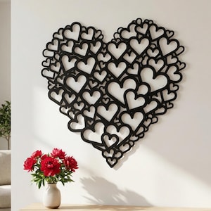 Puede incluir: Decoración de pared en forma de corazón negro, compuesta por numerosos corazones más pequeños. El diseño está montado en una pared blanca. Un jarrón con flores rojas se encuentra sobre una superficie de madera.