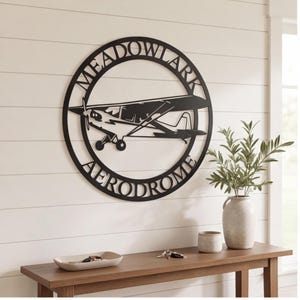 May include: A circular black metal wall sign featuring a silhouette of a small propeller airplane in the center. The text MEADOWLARK is arched along the top inner edge of the circle, and the word AERODROME is arched along the bottom inner edge.