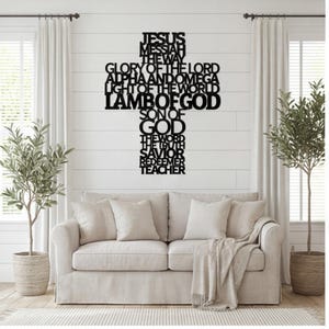 May include: A black metal wall art piece shaped like a cross, featuring stacked text including JESUS, MESSIAH, THE WAY, GLORY OF THE LORD, ALPHA AND OMEGA, LIGHT OF THE WORLD, LAMB OF GOD, SON OF GOD, THE WORD, THE TRUTH, SAVIOR, REDEEMER, and TEACHER.