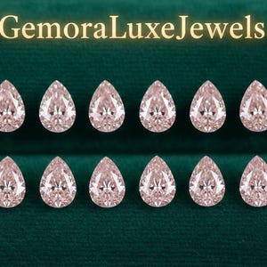 May include: Ten pear-shaped, light pink gemstones are arranged in two rows on a dark green velvet surface. The gemstones are faceted, reflecting light. The text "GemoraLuxeJewels" is displayed in gold lettering at the top.
