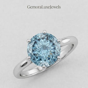 May include: A silver ring featuring a round, faceted light blue gemstone held by six prongs. The band is smooth and polished. The text GemoraLuxeJewels is visible at the top of the image. The design is centered on a plain, light gray background.
