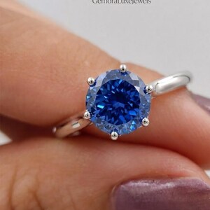 Può includere: Un anello in argento con una pietra preziosa blu rotonda dal taglio brillante, fissata da sei griffe. La fascia è liscia e lucida. Il testo GemoraLuxeJewels è visibile nella parte superiore dell'immagine, indicando il marchio.