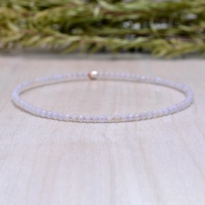 Pode incluir: Uma delicada pulseira de contas roxas claras. A pulseira é feita de pequenas contas facetadas unidas. As contas têm um brilho sutil e são amarradas em um cordão fino e flexível. A pulseira é mostrada em uma superfície de madeira clara.