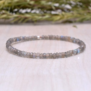 May include: A delicate bracelet composed of small, faceted, gray-toned beads. The beads have subtle blue and iridescent flashes. The bracelet is strung on a thin, elastic band. The jewelry is displayed on a light-colored surface.