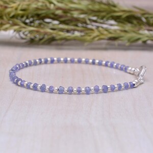 May include: A delicate bracelet featuring small, light purple tanzanite beads interspersed with silver-colored beads. The bracelet is secured with a silver-colored clasp. The jewelry is displayed on a light-colored wooden surface.