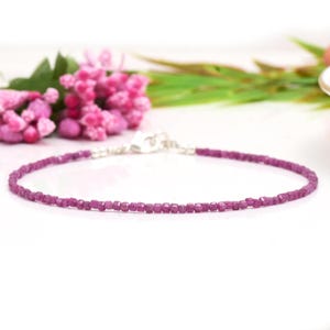 Pode incluir: Uma delicada pulseira magenta com pequenas pedras preciosas de corte quadrado. A pulseira tem um fecho prateado e é apresentada sobre um fundo branco com flores cor-de-rosa e folhagem verde.