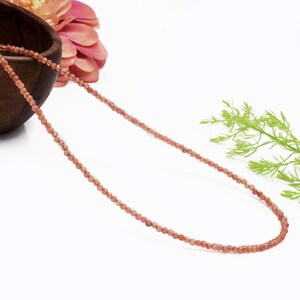 Puede incluir: Delicado collar con pequeñas cuentas redondas de color melocotón. Las cuentas están ensartadas para formar un hilo largo. El collar se exhibe sobre una superficie blanca, con un cuenco de madera y una flor rosa en el fondo.