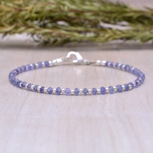 May include: A delicate bracelet featuring small, round, light purple beads interspersed with silver-colored beads. The bracelet is strung on a thin, flexible wire and has a silver clasp. The background is a light wood surface with green foliage.