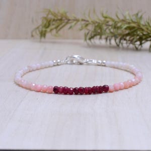 Puede incluir: Una pulsera delicada con un degradado de cuentas rosas y blancas, que conduce a una fila de cuentas de color rojo intenso. Tiene un cierre plateado. La joya se exhibe sobre una superficie de madera clara. Las cuentas miden unos 0,3 cm de diámetro.