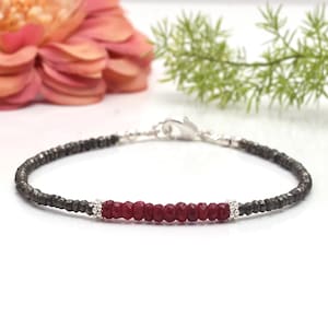 May include: A delicate bracelet featuring a central row of faceted, deep red gemstones, flanked by dark gray, metallic beads. The bracelet is secured with a silver clasp. The background includes a blurred pink flower and green foliage.