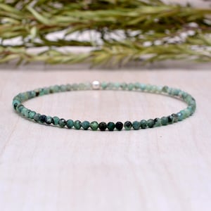 Op de afbeelding: Een delicate armband met kleine, gefacetteerde smaragdgroene kralen in verschillende groentinten. De kralen zijn aan elkaar geregen, waardoor een flexibele band ontstaat. Een kleine, zilverkleurige kraal is zichtbaar. De armband wordt op een licht oppervlak gepresenteerd.