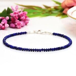 Op de afbeelding: Een delicate armband met kleine, gefacetteerde, diepblauwe kralen. De kralen zijn geregen met een zilverkleurige sluiting. De armband wordt gepresenteerd op een wit oppervlak, met roze bloemen en groene bladeren op de achtergrond.