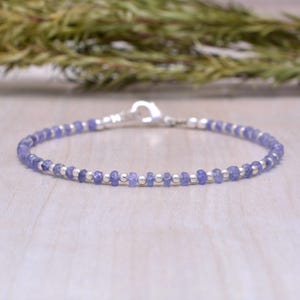 May include: A delicate bracelet featuring small, round, light purple beads interspersed with silver-colored beads. The bracelet is strung on a thin, flexible wire and secured with a silver-colored clasp. The bracelet is approximately 7 inches long.