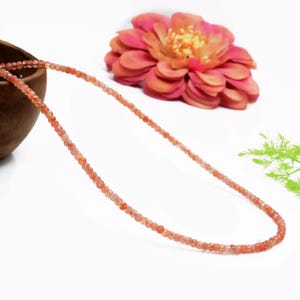 Puede incluir: Un delicado collar compuesto por pequeñas cuentas redondas de color naranja. El collar se exhibe sobre una superficie blanca, con una gran flor rosa y naranja de múltiples capas en el fondo. Un cuenco de madera es parcialmente visible.