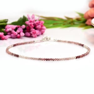May include: Delicate beaded bracelet with a gradient of pink and burgundy beads. The bracelet is strung on a thin, silver chain and is displayed on a white surface with pink flowers in the background.