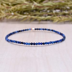 Może przedstawiać: Delikatna bransoletka złożona z małych, okrągłych, ciemnoniebieskich koralików. Koraliki są nanizane, tworząc elastyczną opaskę. Bransoletka jest prezentowana na jasnej drewnianej powierzchni z rozmytym zielonym tłem.