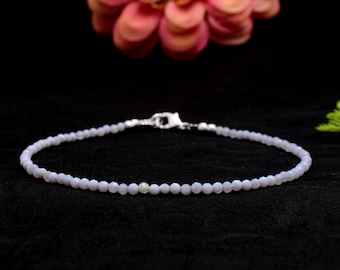 Puede incluir: Una delicada pulsera de cuentas azul claro con un cierre plateado. La pulsera está compuesta por pequeñas cuentas redondas y se muestra sobre una superficie negra. Una flor rosa está en el fondo.