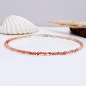 Può includere: Una delicata collana composta da piccole perline sfaccettate arancione-rossastre. Le perline sono infilate con una chiusura argentata. La collana è esposta su una superficie bianca, con una conchiglia e una pianta secca sullo sfondo.