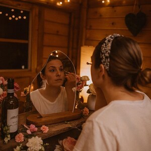 May include: A woman applying makeup, reflected in an unusual mirror. The mirror is on a wooden table decorated with flowers and a wine bottle. The scene is lit by fairy lights, creating a warm and inviting atmosphere.