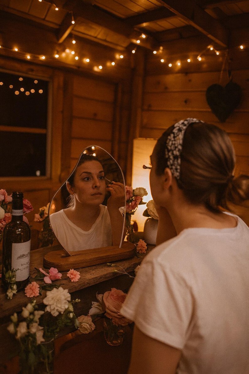 May include: A woman applying makeup, reflected in an unusual mirror. The mirror is on a wooden table decorated with flowers and a wine bottle. The scene is lit by fairy lights, creating a warm and inviting atmosphere.