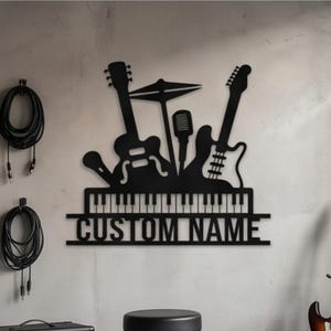 May include: A black metal wall art piece featuring silhouettes of two guitars, a microphone, and cymbals positioned above a piano keyboard design. Below the keyboard, the text CUSTOM NAME is cut out in a bold, sans-serif font.