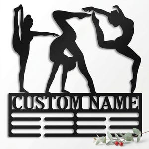 Puede incluir: Arte de pared de metal negro con tres figuras de gimnastas estilizadas en poses dinámicas. Debajo de las figuras hay un panel rectangular con el texto "CUSTOM NAME" y tres barras horizontales. Una ramita de vegetación con bayas rojas en la parte inferior.