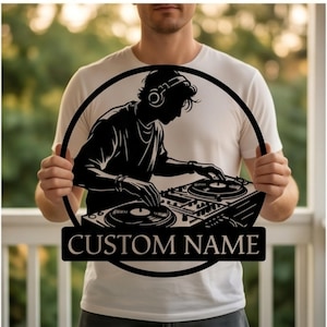 Puede incluir: Un letrero de pared circular negro que presenta la silueta de un DJ con auriculares operando una mesa de mezclas. Debajo del marco circular, una pancarta rectangular contiene el texto CUSTOM NAME en una fuente serif, todo en color negro.