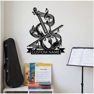 May include: A black metal wall art piece featuring a violin, a bow, musical staff lines, and a treble clef. Below the instrument is a banner with the text CUSTOM NAME. The design is a flat, silhouette-style cutout intended for wall mounting.