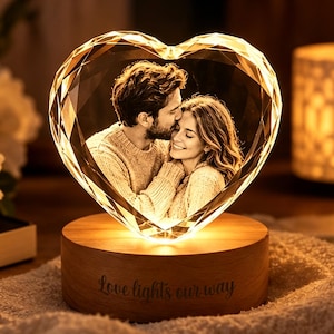 Pode incluir: Um bloco de cristal em forma de coração com uma imagem gravada a laser em 3D de um casal. O cristal assenta numa base de madeira redonda com o texto gravado "Love lights our way" e emite um brilho interno quente que ilumina o retrato.