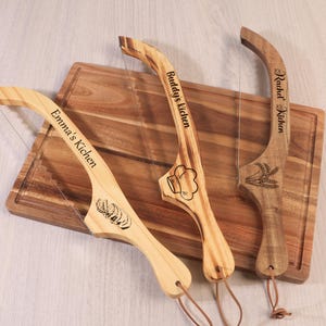 Könnte beinhalten: Drei Käseschneider aus Holz mit Drahtklingen liegen auf einem Holzschneidebrett. Jeder Schneider ist mit einem Namen und einer Lebensmittelillustration graviert. Die Schneider haben Lederschnurschlaufen am Boden.