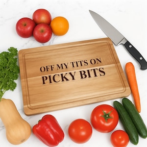 Könnte beinhalten: Ein Holzschneidebrett mit dem Text "OFF MY TITS ON PICKY BITS" ist von frischem Gemüse umgeben, darunter rote Äpfel, eine Orange, Tomaten, Gurken, eine Karotte und eine rote Paprika. Ein großes Messer ist ebenfalls vorhanden.