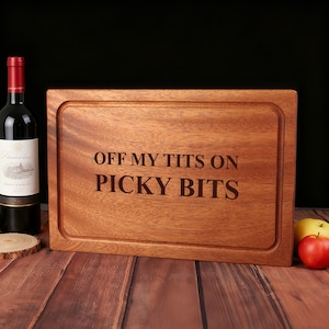 Könnte beinhalten: Ein rechteckiges Holzschneidebrett mit dem eingravierten Text "OFF MY TITS ON PICKY BITS". Links vom Brett befinden sich eine Flasche Rotwein und ein kleiner Holzuntersetzer. Rechts befinden sich ein roter Apfel und ein gelber Apfel.