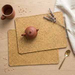 Puede incluir: Dos manteles individuales tejidos con una tetera, una taza y una cuchara sobre una superficie de madera. Los manteles individuales son rectangulares y de color marrón claro natural. Una tetera pequeña y redonda de color marrón está sobre uno de los manteles.