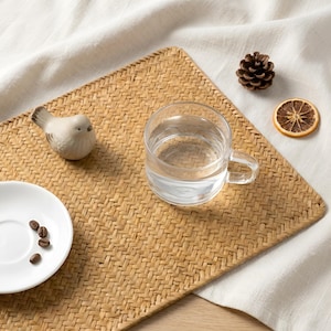 Könnte beinhalten: Ein gewebter, rechteckiger Tischset in Naturtönen mit einer transparenten Glas-Tasse, die mit Wasser gefüllt ist. Ein kleiner Keramikvogel, eine weiße Untertasse mit Kaffeebohnen, ein Tannenzapfen und eine getrocknete Orangenscheibe sind auf der Matte angeordnet.