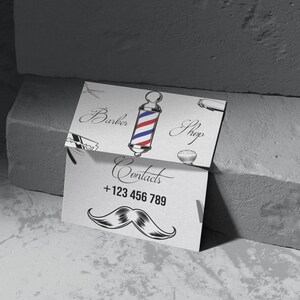 Może przedstawiać: Dwie wizytówki dla zakładu fryzjerskiego. Górna karta zawiera ilustrację słupa fryzjerskiego ze słowami "Barber Shop". Dolna karta przedstawia grafikę wąsów i dane kontaktowe: +123 456 789.