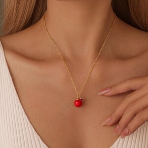 以下が含まれることがあります： 赤いリンゴのペンダントが付いたゴールドカラーのネックレス。リンゴのペンダントには小さな赤い宝石とゴールドカラーの王冠が付いています。ネックレスは人の首に着用されています。