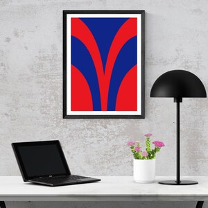 May include: A framed abstract art print with a bold design of red and blue curved shapes. The artwork is displayed on a wall above a desk with a laptop, a black desk lamp, and a small white pot of pink flowers.