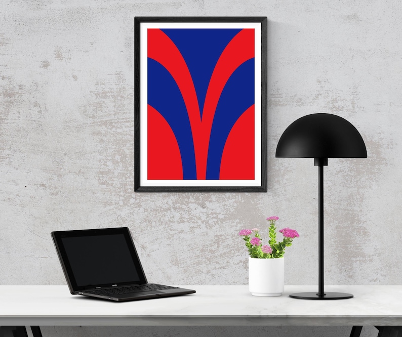 May include: A framed abstract art print with a bold design of red and blue curved shapes. The artwork is displayed on a wall above a desk with a laptop, a black desk lamp, and a small white pot of pink flowers.