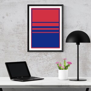 May include: A framed abstract art print with a red and blue colour block design. The artwork features a red upper section, blue lower section, and three horizontal red stripes. A black desk lamp, laptop, and small potted flowers are also visible.