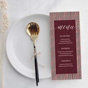 May include: A white plate with a gold fork and black handle, and a menu card with a claret background and white text. The menu card lists starters, entrees, and desserts.