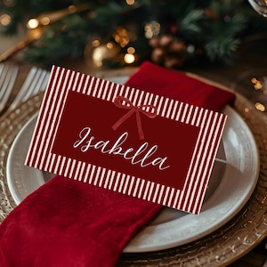 Peut inclure: Une carte de place de couleur bordeaux avec des rayures verticales blanches et le nom "Isabella" en écriture élégante. Un petit nœud orne le dessus. La carte repose sur une serviette rouge, une assiette blanche et un chargeur doré, avec un fond festif.
