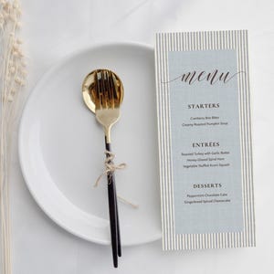 May include: A white plate holds a gold and black fork and spoon tied with twine. Beside it, a menu card features a light blue center with white and gray vertical stripes, listing starters, entrees, and desserts in black text.