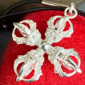May include: A silver Vajra pendant with detailed craftsmanship, comprising a central sphere and four symmetrical prongs. The pendant hangs from a black cord, set against a vivid red backdrop. This spiritual item is a symbol of enlightenment.