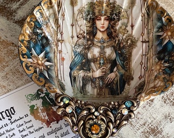 Può includere: Una conchiglia decorativa con un designo dorato e blu che presenta una donna con una corona che tiene una spada. Il testo "VIRGO" è scritto sulla conchiglia. Il testo sotto la conchiglia dice "25 agosto - 22 settembre, Simbolo: Vergine, Colore: Marrone, Elemento: Terra, Erba: Finocchio, Pianeta: Mercurio, Pietra: Zaffiro blu, Vergine, la pratica e l'analitica..."