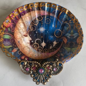 May include: A decorative seashell dish with a blue and gold crescent moon design. The moon has a face and is surrounded by stars. The dish has a gold rim and a jeweled accent.