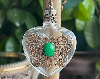 May include: A heart-shaped glass pendant necklace with a butterfly design. The pendant features a bronze-colored filigree butterfly with a green gemstone center. The necklace has a bronze-colored chain and a decorative top.