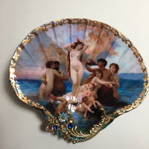 May include: A decorative seashell featuring a painted image of Botticelli's "The Birth of Venus." The shell has a gold-painted edge and is adorned with a gold-toned filigree and blue and pink gemstones. The artwork depicts figures in a sea setting.